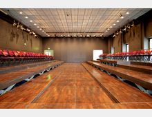 Ein moderner Veranstaltungsraum mit einem hölzernen Laufsteg in der Mitte, flankiert von roten Klappstühlen auf erhöhten Tribünen. Der Raum hat eine abgehängte Decke mit Scheinwerfern und schwarze Wände, die schallisoliert wirken.