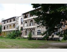 Zweistöckiges, historisches Fachwerkgebäude mit auffälligen Verzierungen an Balkonen und Fenstern, teilweise verfallen. Im Vordergrund eine Wiese mit Bäumen, die das Gebäude teilweise verdecken.