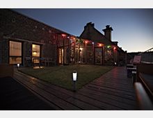 Großer Hof bei Dämmerung mit einem rustikalen Backsteingebäude, großen Glasflächen und einem beleuchteten Gartenbereich. Der Holzdeck-Boden führt zu einem Rasen, umrahmt von bunten Lichterketten und Außenmöbeln.