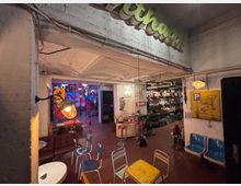 Ein industriell gestalteter Innenraum mit weißen Ziegelwänden, freiliegenden Rohren und einer Mischung aus Retro-Möbeln wie bunten Stühlen und Neonlichtern. Im Hintergrund befinden sich eine Bar mit Hockern, eine Bücherwand und bunte Leuchtschrift-Dekorationen.