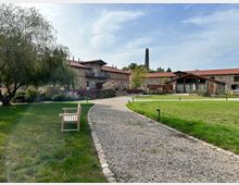 Historisches Anwesen mit Gebäuden aus Ziegelstein und roten Ziegeldächern, umgeben von gepflegten Grünflächen, einem Kiesweg und Blumenbeeten; im Vordergrund eine Holzbank, im Hintergrund ein Schornstein.