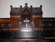 Holzschrank mit gotischen Verzierungen, darunter geschnitzte Gitterelemente und kunstvoll gearbeitete Metallbeschläge, mit einem zentral platzierten Kruzifix auf dem mittleren Regal. Der Schrank steht vor einer schlichten weißen Wand.