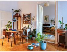 Wohnzimmer mit hellem Holzparkett, einem runden Holztisch und vier Holzstühlen, umgeben von zahlreichen Zimmerpflanzen in Töpfen. Im Hintergrund führt eine offene weiße Tür in ein Arbeitszimmer mit Schreibtisch, Stuhl und Wanddekoration.