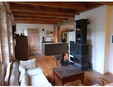 Offener Wohn- und Küchenbereich mit Holzbalkendecke und Holzfußboden, links eine Ziegelwand und eine helle Couch. Rechts steht ein großer schwarzer Kachelofen, im Hintergrund eine moderne Kücheninsel sowie Schränke und eine Tür.