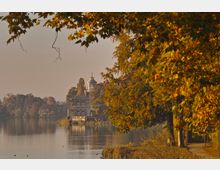 Herbstliche Landschaft mit einem See im Vordergrund, gesäumt von Bäumen mit gelb-orange gefärbtem Laub. Im Hintergrund ein historisches Gebäude mit goldener Kuppel, das direkt am Wasser liegt.