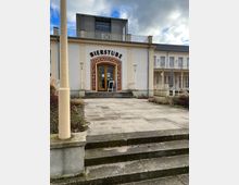 Fassade eines Gebäudes mit der Aufschrift „Bierstube“ über einem braunen Holzeingang mit großen Fenstern, umrahmt von dekorativen roten Verzierungen. Davor führt eine Steintreppe und ein gepflasterter Platz hinauf, umgeben von niedrigem Bewuchs; seitlich ist ein helles, mehrstöckiges Gebäude mit Balkonen sichtbar.