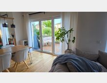 Heller Wohn- und Essbereich mit Holzboden, Esstisch mit Polsterstühlen, Sofa und großer Zimmerpflanze. Eine breite Glas-Schiebetür mit weißen Vorhängen führt auf eine Holzterrasse mit Sitzmöbeln und Blick in einen grünen Garten.