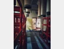 Industriegang in einer technischen Anlage mit roten Metallgeländern und grauem Boden, darüber mehrere große, zylindrische Lüftungs- oder Filterrohre. Am Ende des Ganges stehen schwere weiße Stahltüren mit Handrädern und gelb-schwarzen Warnmarkierungen an den Schwellen.