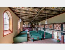 Innenraum einer historischen Kirche mit grün gestrichenen Holzbänken, seitlichen Emporen mit floralen Verzierungen und einem bemalten Holzplafond. Große Bogenfenster lassen Tageslicht herein, und im Hintergrund ist eine kleine Orgel zu sehen.