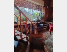 Wohnzimmer mit Holztreppe und großen Fenstern, die von Jalousien bedeckt sind. Im Raum befinden sich Pflanzen, gemütliche Sitzecken mit Kissen, Holzmöbel und ein Teppich auf Holzboden.