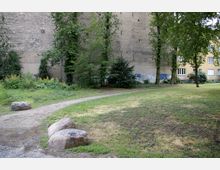 Kleine Grünfläche mit einem schmalen Kiesweg, umgeben von Gras und vereinzelten Bäumen. Im Hintergrund eine alte, beige Hauswand mit Graffiti sowie ein angrenzendes Wohngebäude mit Fenstern.