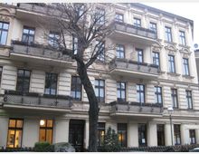 Mehrstöckiges Wohngebäude im Stil der Gründerzeit mit hellen Fassaden, dekorativen Stuckelementen und Balkonen mit schmiedeeisernen Geländern; vor dem Gebäude steht ein kahler Baum.