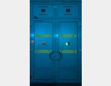 Zweiflügelige Metalltür in blauem Farbton mit gelben Streifen und kleinen rechteckigen Fenstern mit Gittern, ausgestattet mit runden Griffen in Edelstahloptik. Beleuchtung erzeugt eine futuristische Wirkung, die Umgebung ist dunkel.