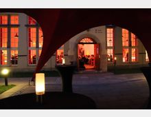 Außenansicht eines Gebäudes mit großen Fenstern, durch die ein beleuchteter Veranstaltungsraum in warmem Rot sichtbar ist. Im Vordergrund befinden sich Stehtische mit Kerzenlichtern und ein rotes Zeltdach, das einen Teil des Blicks rahmt.