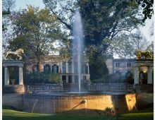 Ein klassizistischer Brunnen mit steinernen Löwenstatuen auf Säulen, flankiert von einem großen, historischen Gebäude im Hintergrund. Rundherum befinden sich Bäume und gepflegtes Grün, die eine parkähnliche Umgebung andeuten.