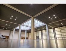 Großer, moderner Innenraum mit hohen Decken, mehreren weißen Säulen und großen, deckenhohen, milchglasähnlichen Fenstern. Der Boden ist glatt und reflektierend, und eine Lichtkuppel aus Glas in der Decke sorgt für zusätzliches Tageslicht.