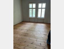 Ein leerer Raum mit frisch abgeschliffenem Holzboden und drei großen, weißen Fenstern mit Sprossen, die nach außen auf eine grüne Umgebung blicken. Die Wände sind schlicht verputzt, und an der linken Wand befinden sich Steckdosen.