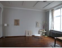 Ein minimalistisch eingerichteter Raum mit Holzboden, einer grauen Wand und großen Fenstern mit weißen Vorhängen, die natürliches Licht einlassen. An der Wand hängen zwei Bilder, und ein kleiner Tisch mit einer Vase, die Zweige enthält, steht in einer Ecke.