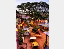 Eine urbane Außenterrasse mit Holzboden, ausgestattet mit Holztischen und Stühlen, umgeben von Pflanzen und kleinen, bunt gestalteten Containern. Im Hintergrund befindet sich ein großer Baum, Backsteinbögen an einer Hauswand und eine beleuchtete Stadtlandschaft.