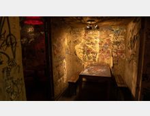 Ein kleiner, düster beleuchteter Raum mit rustikalem Holztisch und zwei einfachen Holzbänken, umgeben von Wänden, die vollständig mit farbigem Graffiti und Kritzeleien bedeckt sind. Ein rotes Lampenschirmlicht und eine Deckenleuchte sorgen für warmes, schwaches Licht.