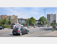Straßenszene mit einem modernen Einkaufszentrum im Hintergrund, das durch große Glasfassaden und säulenartige Strukturen geprägt ist. Im Vordergrund fahren Autos auf einer mehrspurigen Straße, daneben stehen mehrstöckige Wohngebäude und Bäume.