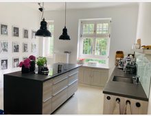 Moderne Küche mit weiß gestrichenen Wänden, einer mittigen Kochinsel mit schwarzer Arbeitsplatte, zwei schwarzen Hängeleuchten und mehreren Schwarz-Weiß-Fotografien an der Wand. Große Fenster mit Sprossenrahmen lassen Tageslicht herein, während Geräte und Utensilien auf den Arbeitsflächen verteilt sind.