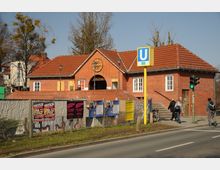Backstein-Bahnhofsgebäude mit rotem Ziegeldach, darüber eine Uhr und der Schriftzug „Kieler Platz“. Im Vordergrund ein U-Bahn-Schild der Linie U3, eine Straße, ein Zaun mit Plakaten und ein Fußgängerüberweg mit Ampel.