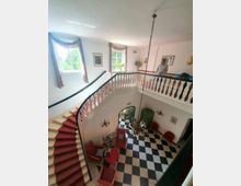 Blick in ein zweistöckiges Treppenhaus mit einer geschwungenen roten Treppe und einem schwarz-weiß karierten Boden. Im unteren Bereich sind rote Stühle und ein Tisch angeordnet, über denen ein Kronleuchter hängt; oben verläuft eine Galerie mit geschnitztem Geländer, und die Wände sind mit Bildern dekoriert.