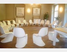 Ein heller Versammlungsraum mit einem Kreis aus weißen, modern geformten Stühlen auf einem Holzboden. Im Hintergrund befinden sich weiße Schränke, eine helle Wand und Pflanzen in weißen Blumentöpfen nahe großen Fenstern, durch die Tageslicht einfällt.