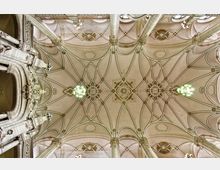 Blick auf eine reich verzierte, symmetrische Gewölbedecke in neogotischem Stil mit filigranen Mustern und floralen Ornamenten, getragen von verzierten Säulen. Im Zentrum der Decke und an den Seiten befinden sich Kronleuchter, die diffuses Licht spenden.