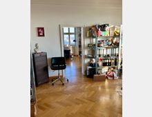 Ein Arbeitszimmer mit hellem Parkettboden, einem schwarzen Bürostuhl und offenen Regalen voller Akten und Bücher. Im Hintergrund führt eine Tür zu einem weiteren Raum mit einem Fenster, durch das eine Stadtlandschaft erkennbar ist.