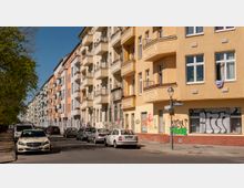 Straßenszene mit mehrstöckigen Wohnhäusern in gelben und pastellfarbenen Tönen. Geparkte Autos säumen die Straße, einige Fassaden zeigen Graffiti, und ein Straßenlaternenpfahl steht an einer Ecke.