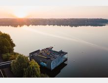 Ein modernes Haus auf Stelzen, das auf einem ruhigen See schwimmt, mit einer begrünten Dachterrasse und Sonnenliegen, umgeben von Bäumen und einem Steg, während im Hintergrund eine bewaldete Uferlandschaft in der Morgensonne liegt.