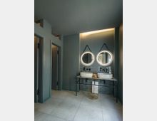 Modernes Badezimmer mit zwei runden, beleuchteten Spiegeln über ovalen Waschbecken auf einem schlichten Metalltisch. Links befinden sich Türen zu getrennten Toiletten, und der Raum hat graue Wände und Bodenfliesen.