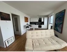 Moderne Wohnküche mit offenem Grundriss, bestehend aus einer weißen Einbauküche mit schwarzen Akzenten, einem kleinen Bartisch mit zwei Hockern und einem beigen Sofa im Vordergrund. Wände sind mit abstrakter Kunst geschmückt, der Boden ist aus Holz, und ein großes Fenster bringt Tageslicht herein.