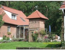 Backsteingebäude mit roten Ziegeldächern, bestehend aus einem Haupthaus mit Dachfenstern und einem kleineren Turmanbau mit Wetterfahne. Im Vordergrund befindet sich eine Gartenfläche mit Blumen, Pflanzen und einer Terrasse; dahinter sind Bäume eines Waldrandes zu sehen.