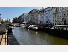 Städtischer Kanal mit mehreren angelegten Booten und historischen Gebäuden auf der rechten Seite, darunter weiße und beige Fassaden mit verzierten Details. Im Hintergrund ist eine Steinbrücke sowie moderne Gebäude zu sehen, umgeben von Bäumen und einem klaren Himmel.