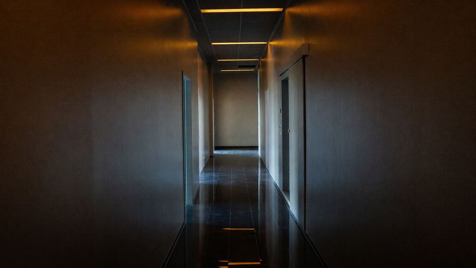 Langer, schmaler Innenflur mit glatten Wänden, dunklem glänzendem Fliesenboden und abgehängter Decke mit rechteckigen Lichtleisten. An beiden Seiten befinden sich mehrere Türen, am Ende des Gangs ist eine helle Wand zu sehen.