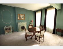 Wohnzimmer mit grünen Wänden, einem kleinen Kamin mit Verzierungen, einem runden Holztisch mit vier Stühlen und hellen Teppichboden. Zwei große, bodenlange Fenster mit beigefarbenen Vorhängen lassen Tageslicht in den Raum, der durch klassische Dekorelemente wie Wandrahmen und ein Landschaftsgemälde ergänzt wird.