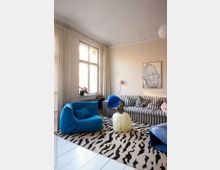 Bright living room with high ceilings, ornate crown molding, and a tall window framed by long curtains. The space features a striped sofa, a blue lounge chair, a zebra-pattern rug, and a small round translucent coffee table beneath a framed wall artwork.