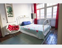 Helles Schlafzimmer mit einem weißen Metallbett und einer bunten Bettdecke, einem modernen weißen Stuhl mit rotem Kissen, einer großen Fensterfront mit roten Vorhängen und Holzboden. An der Wand hängt eine vertikale Bildrolle mit Landschaftsmotiv.