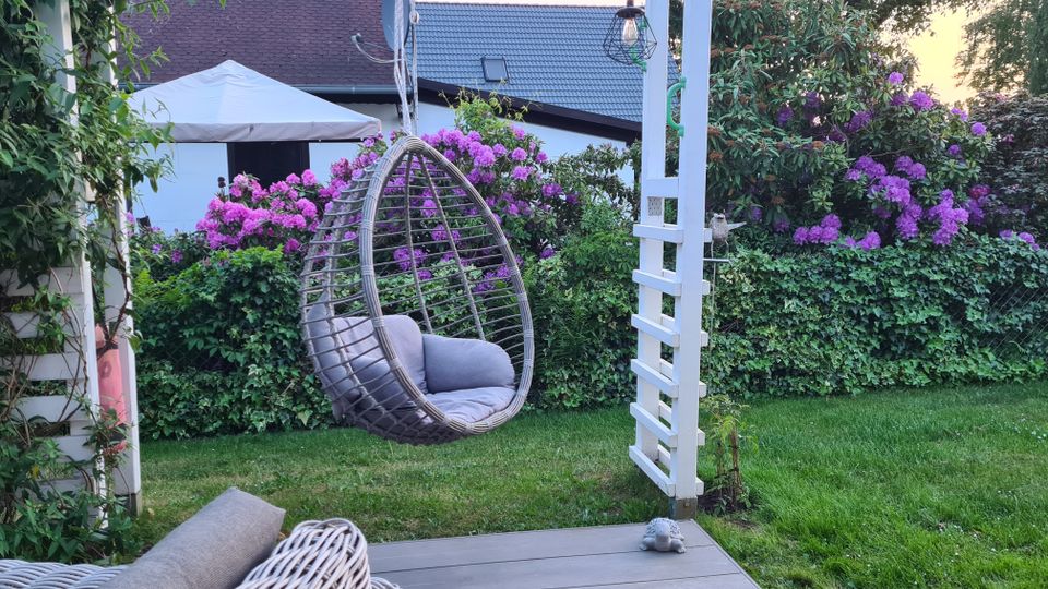 Garten mit einer Terrasse aus Holzplanken, einem Hängesessel aus Rattan und einem weißen Pergola-Gerüst. Im Hintergrund eine grüne Hecke, blühende lila Rhododendren und ein Haus mit hellen Wänden und einem grauen Dach.