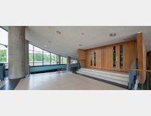 Ein moderner Innenraum mit großen Fenstern, die Blick auf grüne Bäume bieten. Im Vordergrund befinden sich eine Treppe mit Metallgeländer und eine Holzpaneeltür; der Raum ist mit Sichtbetonsäulen und hellen Bodenfliesen gestaltet.