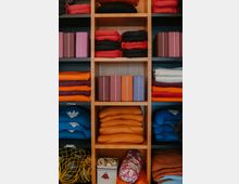 Regal mit mehreren Fächern, gefüllt mit Yogazubehör wie Meditationskissen, Yoga-Blöcken, gefalteten Decken in verschiedenen Farben, Gurtbändern in einer Box und einer gemusterten Nackenrolle. Das Regal besteht aus hellem Holz.