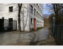 Mehrstöckiges modernes Gebäude mit weißer Fassade und zahlreichen Fenstern; davor ein Gehweg, ein Tor aus Metallgittern und kahle Bäume. Rechts neben dem Gehweg befindet sich ein Zaun, im Hintergrund ist eine parkähnliche Umgebung erkennbar.