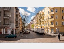 Eine ruhige städtische Straße mit Kopfsteinpflaster, gesäumt von mehrstöckigen Wohngebäuden in gelben und grauen Farbtönen. Geparkte Autos, ein Lieferwagen und wenige Fußgänger sind unter kahlen Bäumen sichtbar, während der Himmel teils bewölkt ist.