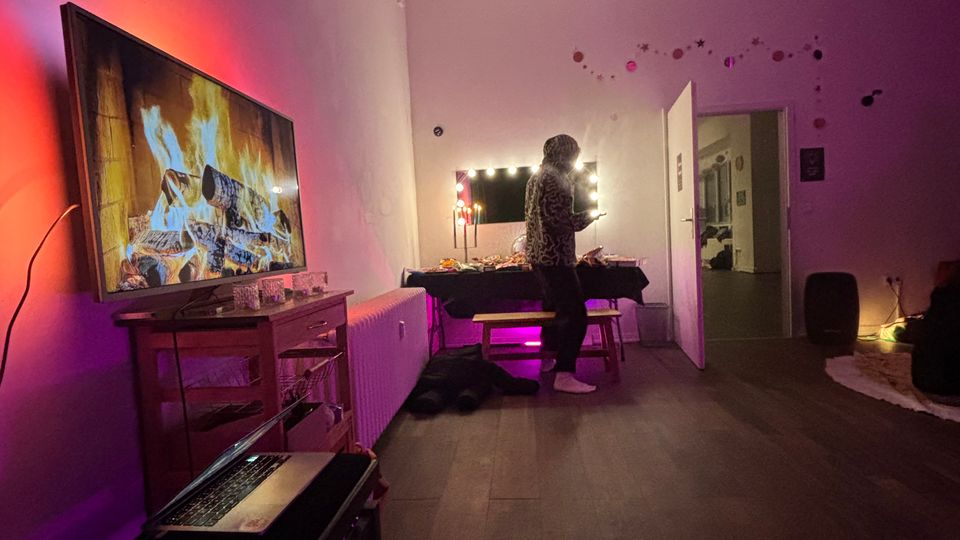 Long, loft-style room with white walls and a high ceiling, featuring purple ambient lighting, a large TV on a low cabinet, and a table with string lights near an open doorway. Acoustic panels are mounted on the ceiling, and the space has wood flooring with minimal furniture.