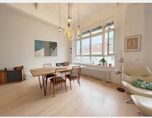 Ein minimalistisch eingerichteter Raum mit hohen Decken, einem rechteckigen Holztisch und vier Stühlen in der Mitte, großen Fenstern mit Blick auf Dächer und mehreren Hängelampen. An den Wänden hängen Bilder, und in einer Ecke steht ein beiges Loungesessel mit einer Stehlampe.