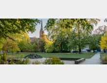 Ein Park mit einer Wiese, Wegen und mehreren Bäumen, im Hintergrund eine rote Backsteinkirche mit einem hohen Turm. Im Vordergrund befindet sich ein kleiner Sandbereich und eine dekorative Metallstruktur.
