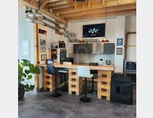 Moderne Küche mit industriellem Design, ausgestattet mit Holz- und Metallakzenten, einer Theke mit hohen Stühlen, einer Spüle, einem Backofen und Hängeschränken. Sichtbare Rohre verlaufen unter einer Holzdecke, und eine digitale Uhr sowie Dekorationselemente schmücken die Wände.